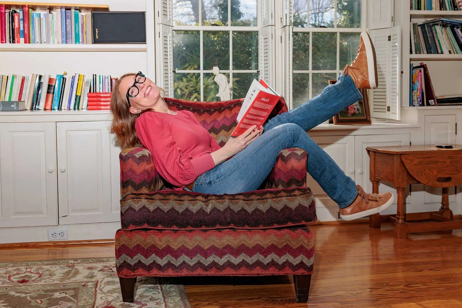 Beth Linker, wearing a bright pink blouse and jeans, lies across an armchair in her living room with one leg kicked up, holding a copy of her book.