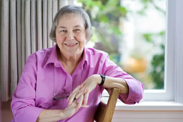 A portrait of Penny Simkin seated on a chair, smiling and looking directly at the camera. She has short gray hair and is wearing a vivid pink blouse.