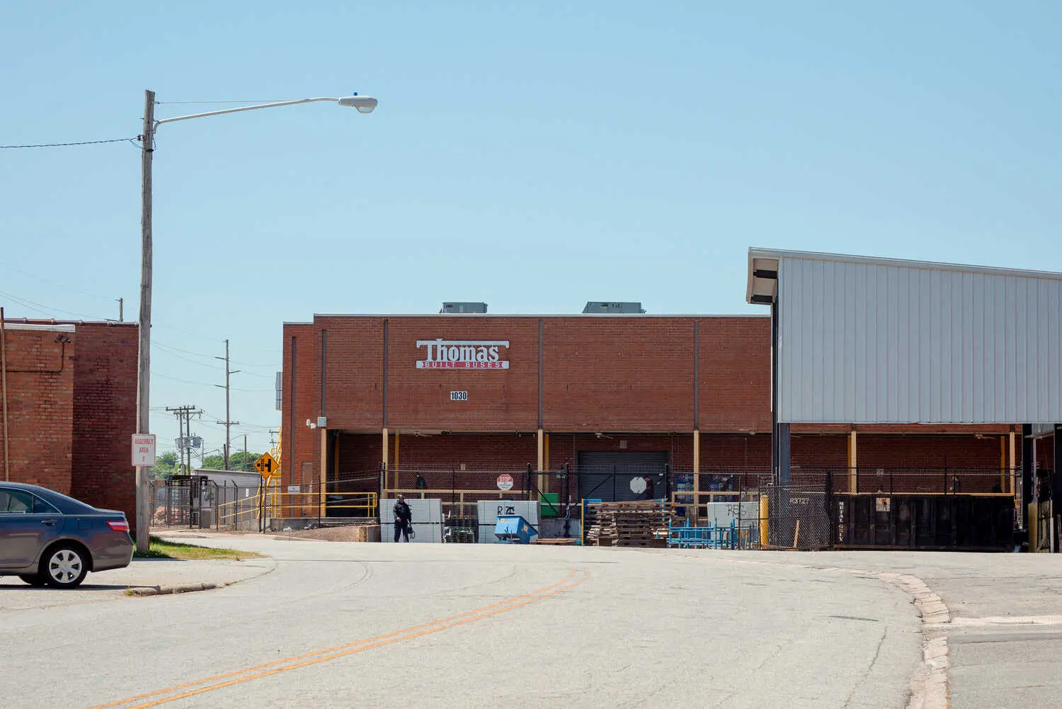 An industrial brick building on a curve in a two-lane road has a sign that says, “Thomas Built Buses.” 