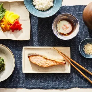 A plate of miso salmon on a navy blue tablecloth with various sides, including a soft-cooked egg and blanched spinach, among others.